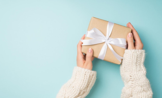 Foto de vista superior en primera persona de manos femeninas en suéter blanco acogedor sosteniendo caja de regalo de papel artesanal con lazo de cinta de raso blanco sobre fondo azul pastel aislado con espacio de copia