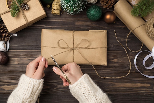 Foto de vista superior en primera persona de manos femeninas en suéter empacando caja de regalo de papel kraft con tijeras de lazo de hilo bolas de oro verde ramitas de pino y herramientas artesanales en un fondo aislado de escritorio de madera oscura