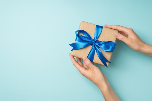 Foto foto de vista superior en primera persona de las manos de la mujer sosteniendo una caja de regalo de papel artesanal con lazo de cinta de raso azul sobre fondo azul pastel aislado con espacio de copia