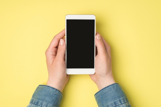 Foto de vista superior en primera persona de las manos de la mujer sosteniendo la pantalla del teléfono inteligente en un fondo amarillo aislado con espacio en blanco