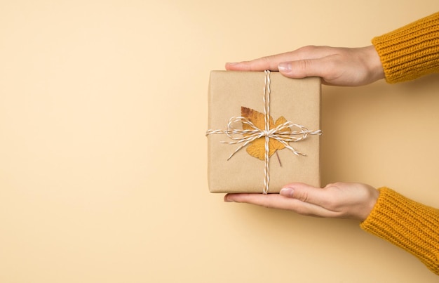 Foto de vista superior en primera persona de manos de mujer en suéter de punto amarillo sosteniendo una elegante caja de regalo de papel artesanal con lazo de hilo y hoja de otoño amarilla sobre fondo beige aislado con espacio vacío