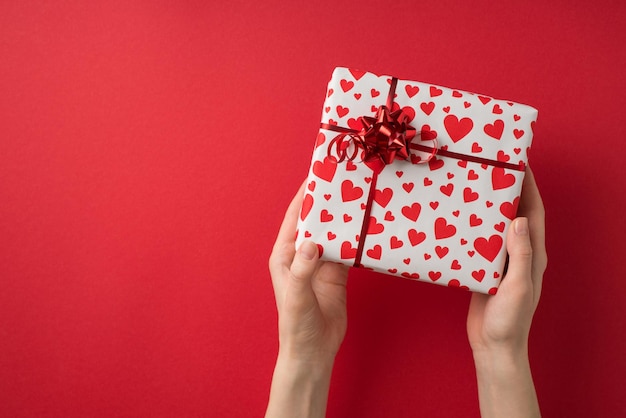 Foto de vista superior en primera persona de las manos de la niña de la decoración del día de san valentín dando una caja de regalo en papel de regalo blanco con patrón de corazón y lazo de estrella roja sobre fondo rojo aislado con espacio de copia