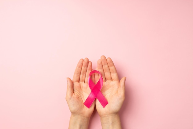 Foto de vista superior en primera persona de las manos de la niña sosteniendo una cinta rosa en las palmas, símbolo de la conciencia del cáncer de mama en un fondo rosa pastel aislado con espacio en blanco