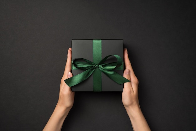 Foto de vista superior en primera persona de manos sosteniendo una caja de regalo negra con lazo de cinta de raso verde sobre fondo negro aislado