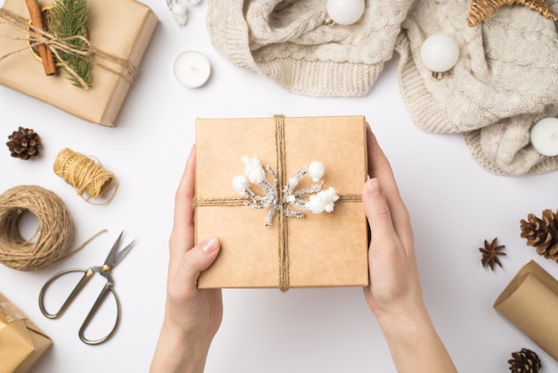 Foto de vista superior en primera persona de manos sosteniendo una caja de regalo de papel artesanal decorada con hilo y rama de nieve sobre bolas de árbol de Navidad suéter y herramientas artesanales sobre fondo blanco aislado