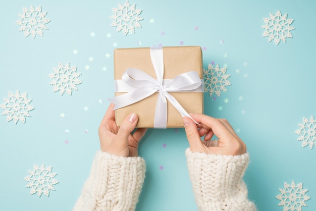 Foto de vista superior en primera persona de manos en suéter blanco desempacando caja de regalo de papel artesanal con lazo de cinta blanca sobre grandes copos de nieve decorativos y confeti brillante sobre fondo azul pastel aislado