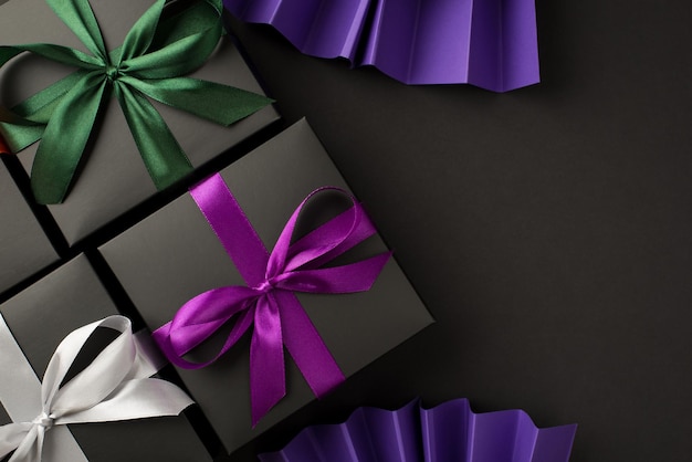 Foto de vista superior de tres cajas de regalo negras con lazos de cinta verde y blanco violeta y abanicos de papel violeta sobre fondo negro aislado con espacio en blanco