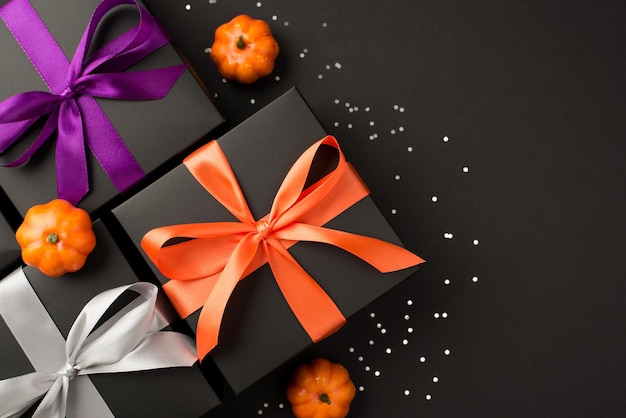 Foto de vista superior de tres cajas de regalo negras con lazos de cinta violeta, naranja y blanco, calabazas pequeñas y lentejuelas brillantes sobre fondo negro aislado con espacio vacío