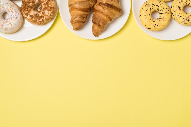 Foto de vista superior de tres platos con donuts y croissants glaseados de diferentes colores sobre fondo amarillo claro aislado con espacio vacío