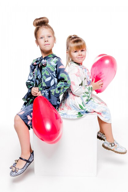 Foto von 2 kleinen Mädchen mit eleganten Kleidern sitzen mit großen Taschen mit herzförmigen Luftballons im Inneren