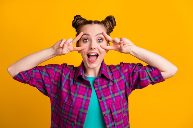 Foto von attraktiver aufgeregter funky Teenager-Dame zwei lustige Brötchen gute Laune überglücklich mit V-Zeichen-Symbol offener Mund sehen Freunde lässiges kariertes Hemd tragen isoliert gelber heller Farbhintergrund