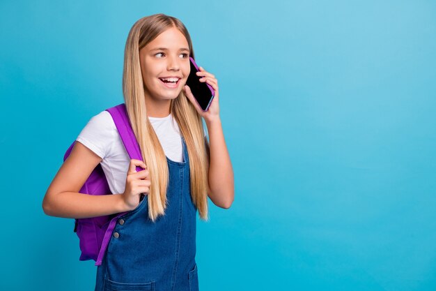 Foto von coolen hübschen Teenager-Mädchen, die leeren Raum suchen, Telefon tragen Tasche Jeanskleid isoliert auf pastellblauem Hintergrund