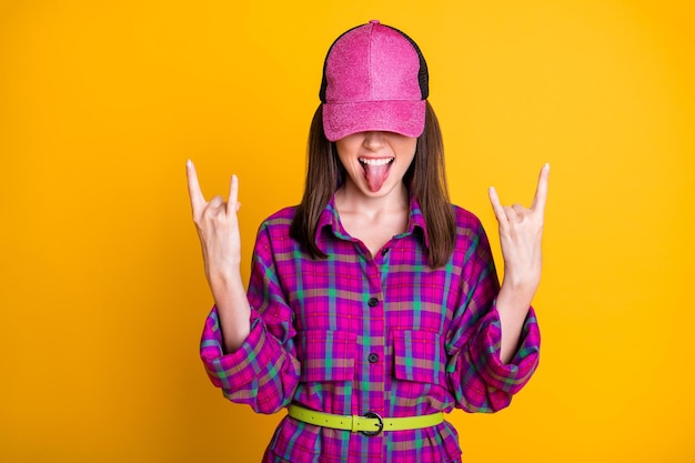 Foto foto von coolen, unhöflichen braunen haaren, mädchen, gesicht, zunge raus, rockgeste, rosa kopfbedeckung, hemdband, isoliert auf gelbem hintergrund