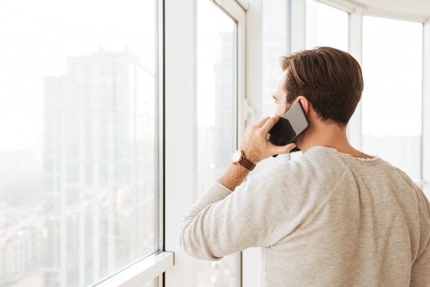 Foto von der Rückseite des kaukasischen Mannes mit kurzen braunen Haaren, die durch Fenster schauen, während auf schwarzem Smartphone sprechen