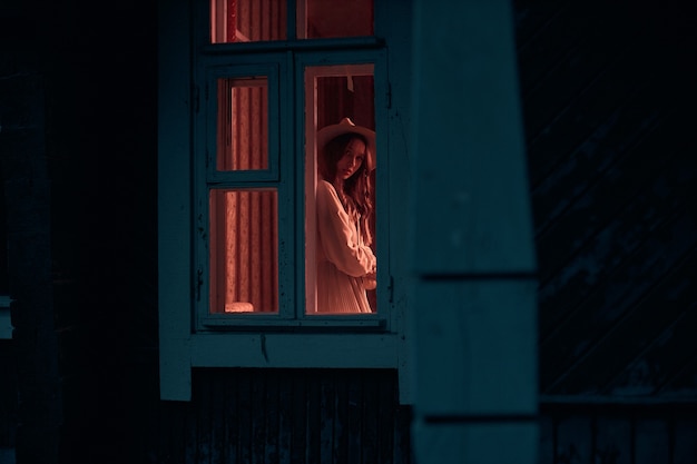 Foto von der Straße zu dem mysteriösen Mädchen, das im Fenster in einem Raum mit den Lichtern auf kreativer Fotografie steht, hochwertiges Foto