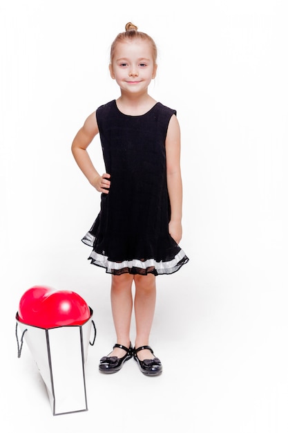 Foto von ein kleines modisches mädchen in einem schwarzen kleid steht neben einem großen paket mit einem herzförmigen ballon im inneren