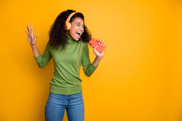 Foto von einem lässigen, fröhlichen, hübschen Jugendlichen, der ins Telefon singt und sich vorstellt, dass es ein Mikrofon ist, der in Jeans-Denim schreit, isolierter gelber, lebhafter Hintergrund