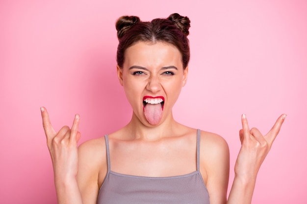 Foto von erstaunlichen jungen Brötchen lustige Brötchen rote Pomade zeigt Finger Hörner, die Zunge heraus Mund verrückte Rocker beim Konzert tragen lässige graue Tank-Top isolierte rosa Farbe