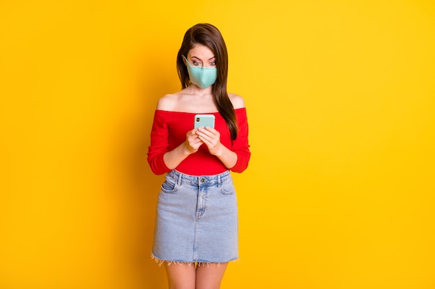 Foto von erstauntem Mädchen in medizinischer Maske mit Smartphone lesen soziales Netzwerk Quarantäne verlängern Nachrichten tragen roten Denim-Jeansrock einzeln auf hell glänzendem Hintergrund