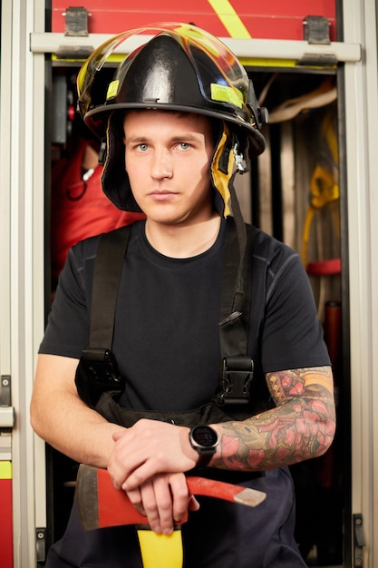 Foto von Feuerwehrmann mit Helm mit Axt gegen Feuerwehrauto Bild von jungem Feuerwehrmann mit Helm im Kopf vor dem Hintergrund eines Feuerwehrautos