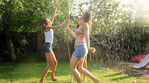 Foto von fröhlichen, fröhlichen Mädchen in nasser Kleidung, die unter Wassergartenschlauch tanzen und springen. Familie, die im Sommer im Freien spielt und Spaß hat