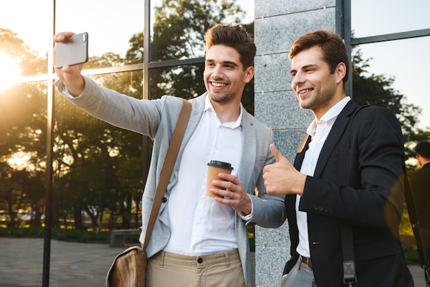 Foto von fröhlichen Geschäftspartnern in Anzügen, die Smartphones für Selfie verwenden, während im Freien nahe Gebäude mit Kaffee zum Mitnehmen stehen