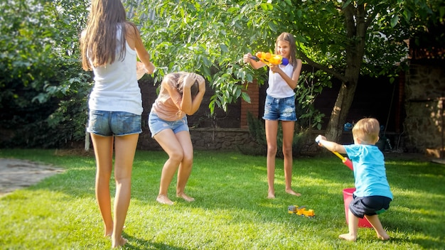 Foto von glücklichen Kindern, die Wasserpistolenkampf im Hausgarten haben. Familie, die im Sommer im Freien spielt und Spaß hat
