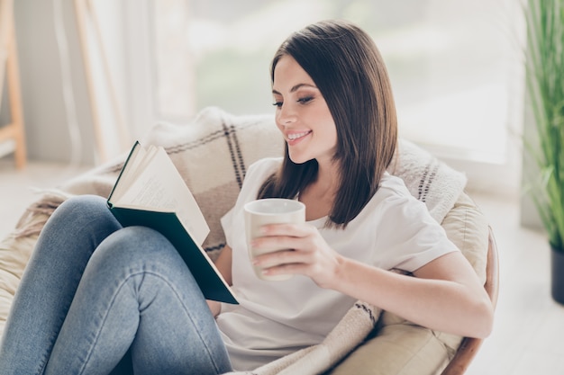 Foto von hübschen jungen Mädchen sitzen Stuhl lesen interessantes Buch halten Teetasse tragen Jeans im Haus drinnen