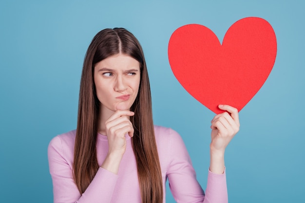 Foto von hübschen Mädchen denken hmm Zweifel sehen große Papierherz-Valentinstag-Gefühle einzeln auf blauem Farbhintergrund aus