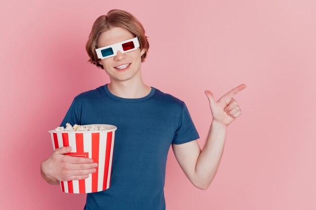 Foto von Kerl halten Popcorn glücklich positives Lächeln tragen 3D-Brille Zeigefinger leeren Raum Ad-Beratung isoliert rosa Farbhintergrund