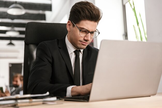 Foto von konzentriertem Geschäftsmann mit Brille, der am Laptop tippt, während er am Schreibtisch im Großraumbüro sitzt