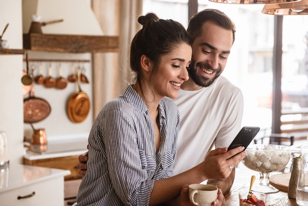 Foto von lächelnden brünetten Paaren, Mann und Frau, die Kaffee trinken und während des Frühstücks in der Küche zu Hause Handy benutzen?