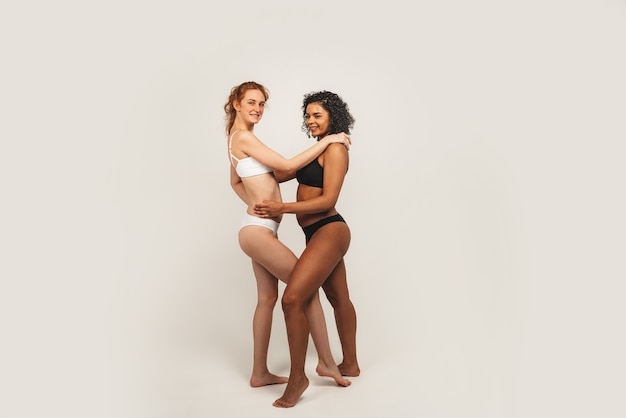 Foto von natürlichen gemischtrassigen Frauen, Körper positiv. Feministische Frauen in Unterwäsche umarmen sich, isoliert auf weißem Hintergrund
