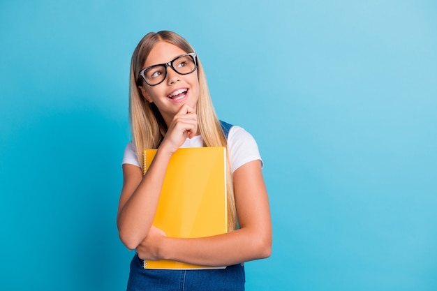 Foto von niedlichen fröhlichen schülermädchen, die denken, dass sie einen leeren raum sehen, tragen eine brille, die ein buch isoliert auf pastellblauem hintergrund hält