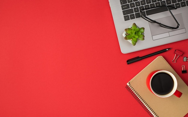 Foto von oben mit Brille, Blumentopf auf Laptop-Mappe, Clips, Stift und roter Tasse Getränk auf Spiralorganisator auf isoliertem rotem Hintergrund mit leerem Raum