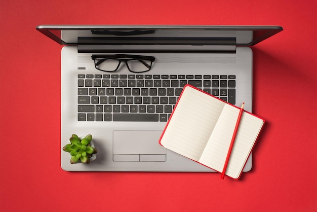 Foto von oben mit Brille, Blumentopf, offener roter Planer und Bleistift auf offenem, grauem Laptop auf isoliertem rotem Hintergrund mit Leerzeichen