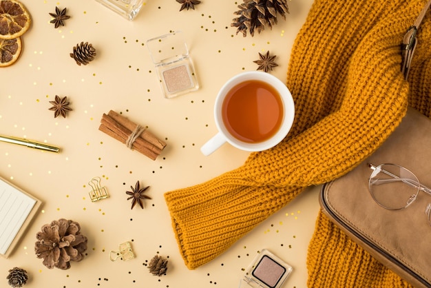 Foto von oben mit gelbem Pullover, Leder, Geldbörse, Klemmen, Stift, Notizbuch, Tasse Tee, Lidschatten, Brille, Tannenzapfen, Anis, getrockneten Zitronenscheiben, Zimt, goldenen Pailletten auf isoliertem, hellbeigem Hintergrund