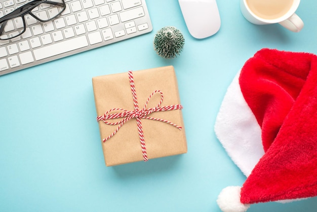 Foto von oben mit rotem Weihnachtsmann-Hut, Brille, Tastatur, Maus, Tasse mit heißem Trinkspielzeug aus Kiefernholz und Bastelpapier-Geschenkbox mit Garnschleife auf isoliertem pastellblauem Hintergrund