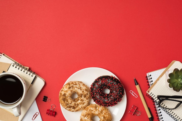 Foto von oben von Arbeitsplatzstapeln von Planern, Brillenbindern, Stiften, Pflanzentellern mit drei verschiedenfarbig bestreuten Donuts und einer Tasse Kaffee auf isoliertem rotem Hintergrund mit Kopierraum