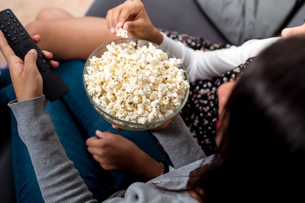 Foto von oben von zwei Mädchen, die auf dem Sofa sitzen, eine von ihnen bedient die Fernbedienung und die andere nimmt Popcorn auf.