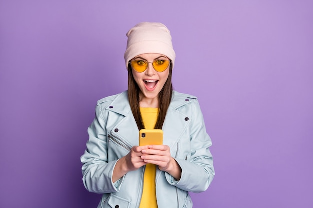 Foto von stilvoller hübscher Dame Straßenkleidung halten Telefon lesen Freunde chatten mit offenem Mund gute Laune tragen Sonnenbrille lässiger Hut blaue moderne Jacke isoliert lila Farbhintergrund