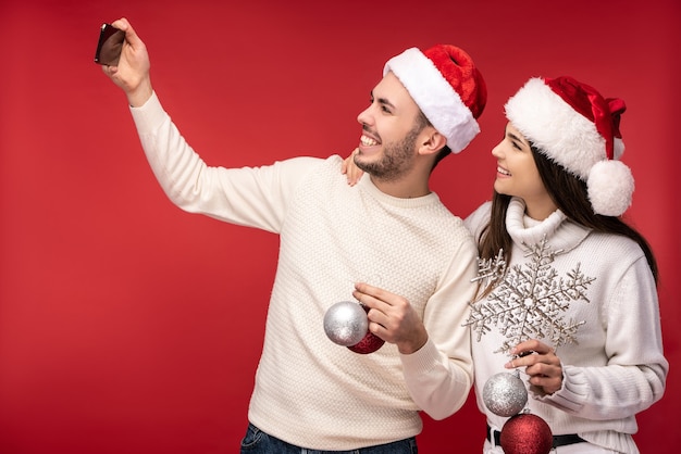 Foto von süßen Paaren in Weihnachtsmützen und mit Weihnachtsspielzeug, sieht glücklich aus. Mann und Frau machen ein Selfie und lächeln, isoliert auf rotem Hintergrund.