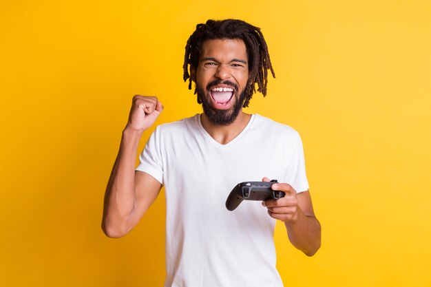 Foto von verrückten schwarzen Kerl halten Joystick Joystick offener Mund heben Arm Faust tragen weißes T-Shirt isoliert gelber Hintergrund