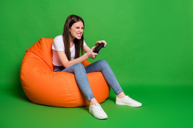 Foto von wütenden lustigen brünetten Haaren junge Frau spielt Playstation tragen T-Shirt Jeans Schuhe sitzen auf Hocker isoliert auf grünem Hintergrund