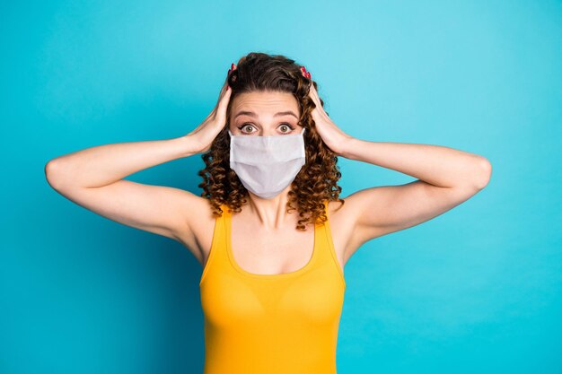 Foto von ziemlich verängstigten Studentenmädchenhänden, die den Kopf starren, tragen Maske gelbes Unterhemd isoliert blauer Farbhintergrund