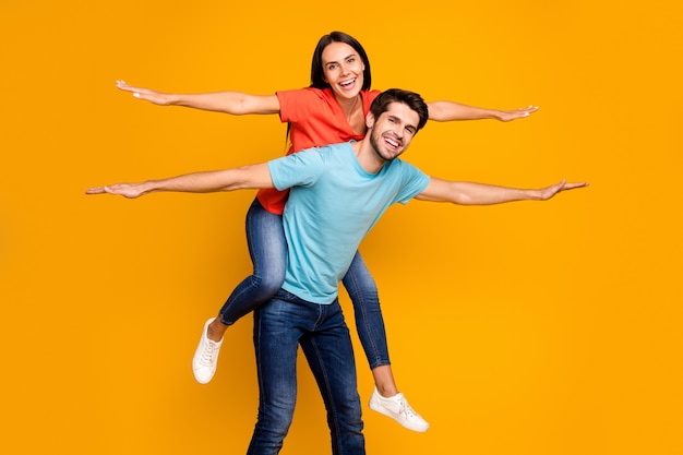 Foto von zwei lustigen Leuten Kerl tragen Dame Huckepack treffen Sommerzeit zusammen verbreiten Hände wie Flügel tragen lässig stilvolle blau orange T-Shirts Jeans isoliert gelbe Farbe Wand