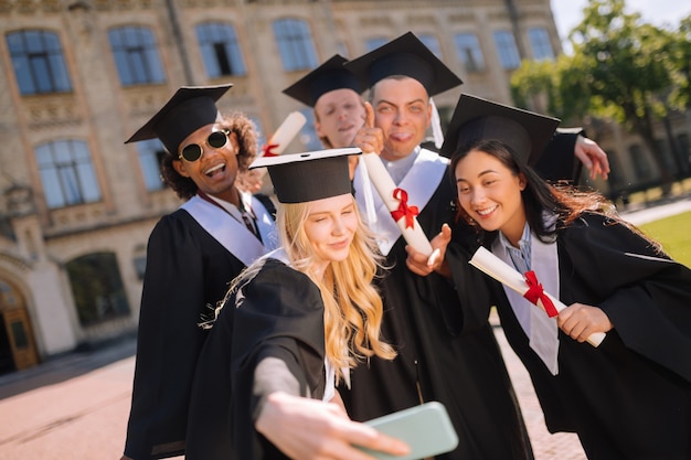 Foto zur Erinnerung. Schönes Mädchen, das nach dem Abschluss auf dem Universitätshof mit ihren Gruppenkameraden fotografiert.