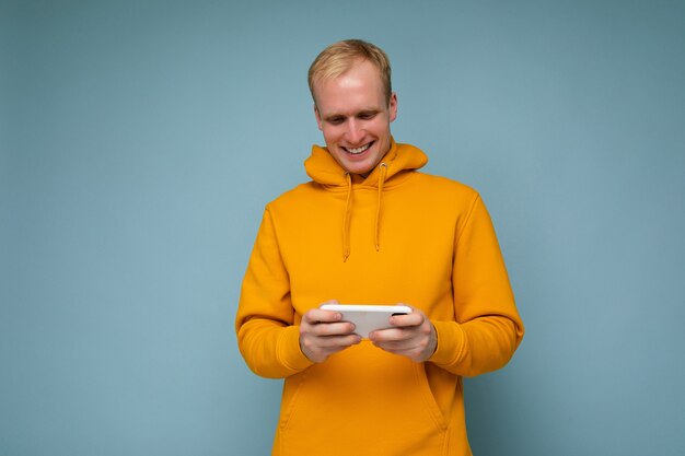 Fotoaufnahme der schönen positiven gut aussehenden jungen blonden männlichen Person, die gelben Kapuzenpulli trägt