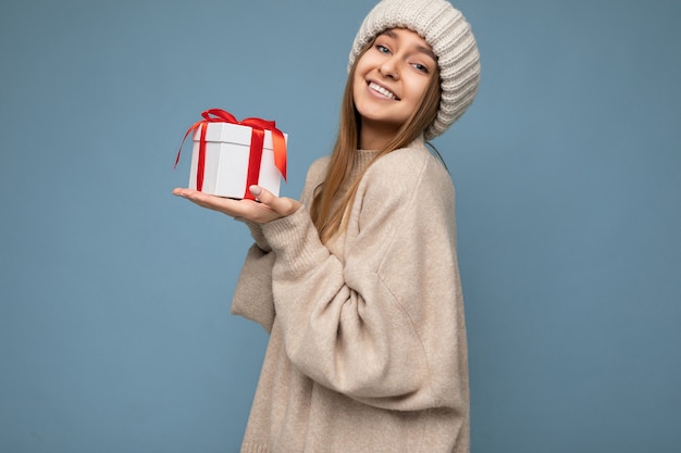 Fotoaufnahme der sexy charmanten glücklichen lächelnden dunkelblonden jungen Frau lokalisiert über blaue Wandwand, die stilvollen Pullover und Wintermütze hält, die weiße Geschenkbox mit rotem Band und hält.