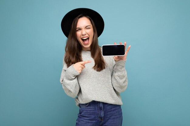 Fotoaufnahme einer schönen jungen Frau mit schwarzem Hut und grauem Pullover, die das Telefon zeigt